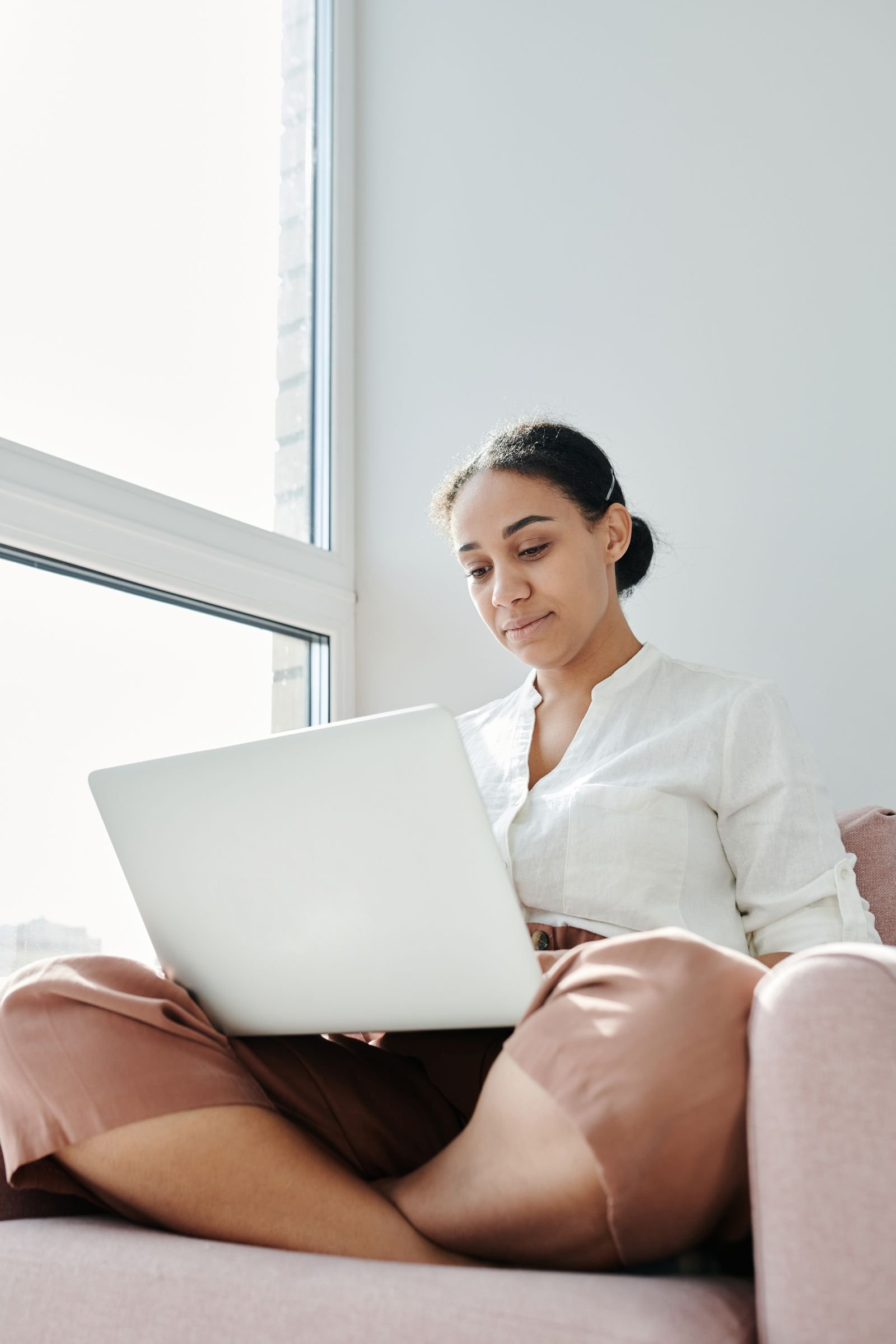 A woman sitting by a window at home, concentrating on a laptop balanced on her lap.