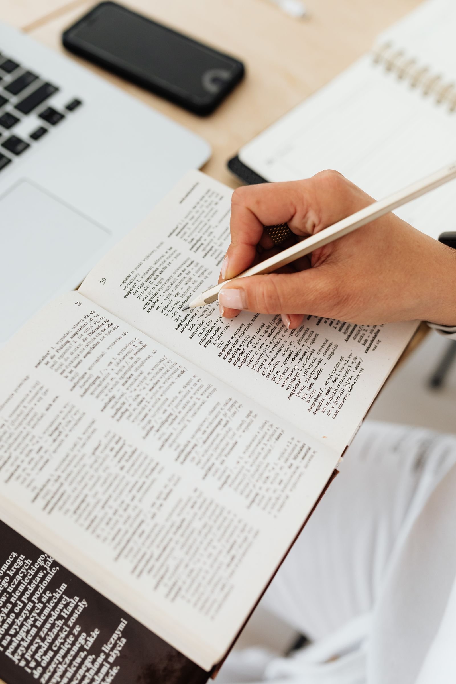 A hand holding a pen over the open pages of a printed dictionary, with a laptop and a notebook on the desk alongside. A hand holding a pen over the open pages of a printed dictionary, with a laptop and a notebook on the desk alongside.