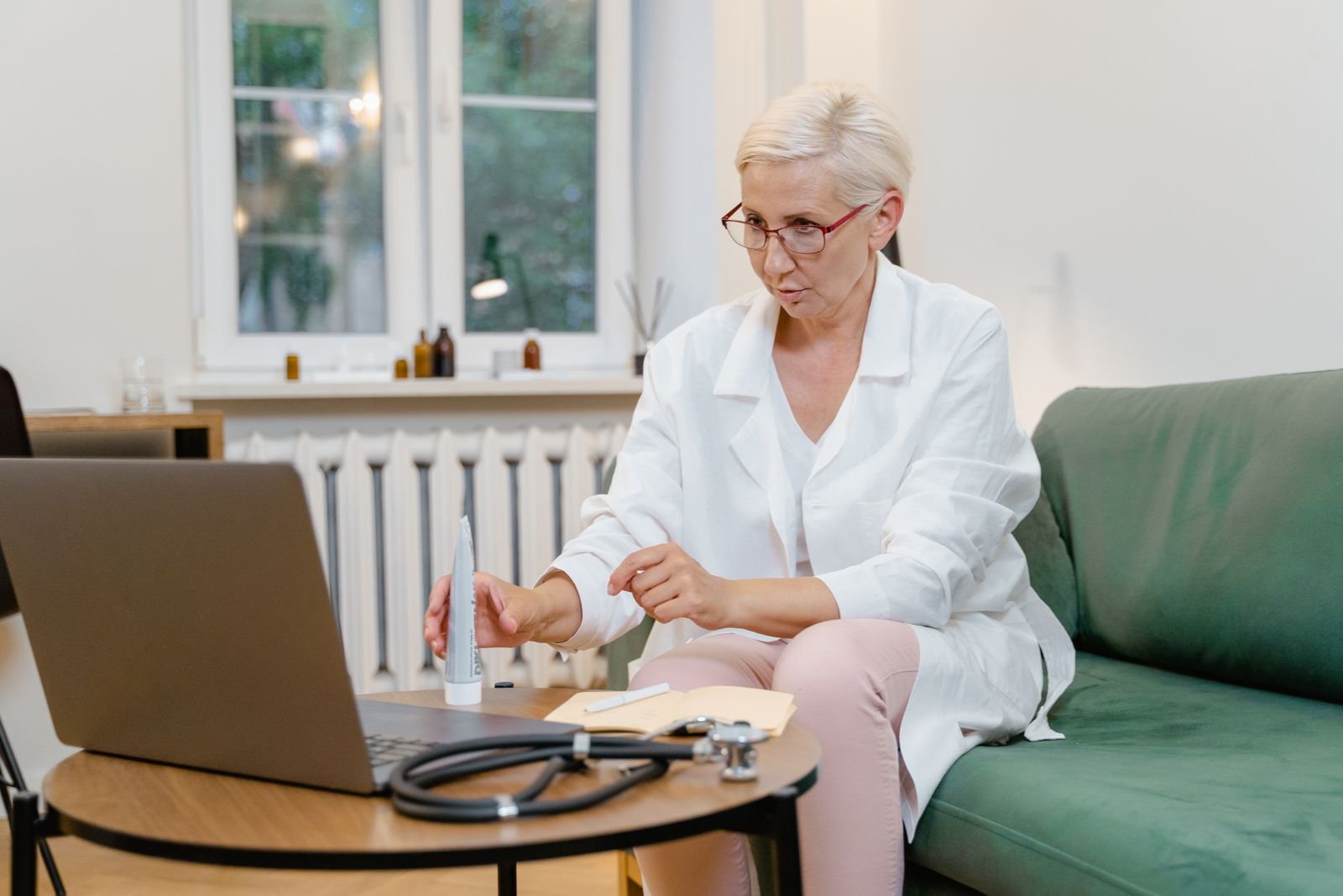 A clinician in a white coat sitting at a low table, consulting a patient through a laptop screen, with a stethoscope and notes beside her. A clinician in a white coat sitting at a low table, consulting a patient through a laptop screen, with a stethoscope and notes beside her.