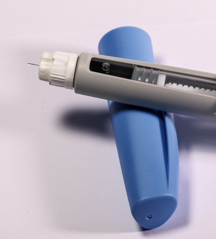 A semaglutide injection pen resting on its blue cap, with a fine needle fitted at the tip, on a plain pale background.